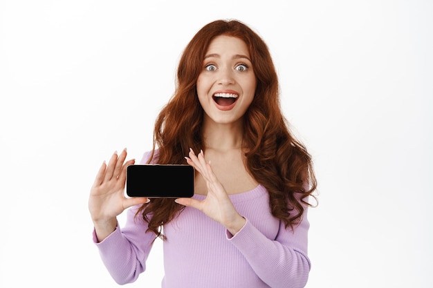 Mulher sorridente e ruiva segurando um smartphone em branco