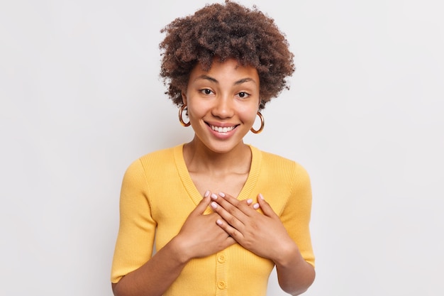 Mulher sorridente e satisfeita pressiona as mãos no peito e expressa gratidão sente-se tocando usa brincos macacão amarelo confortável isolado sobre a parede branca