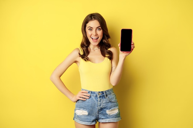 Mulher sorridente elegante mostrando a tela do celular, interface do aplicativo no smartphone, de pé na regata e shorts contra fundo amarelo.