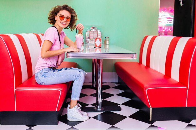 Mulher sorridente elegante no café retrô dos anos 50 sentado à mesa bebendo coquetel de milk-shake com roupa de hipster se divertindo rindo de humor alegre