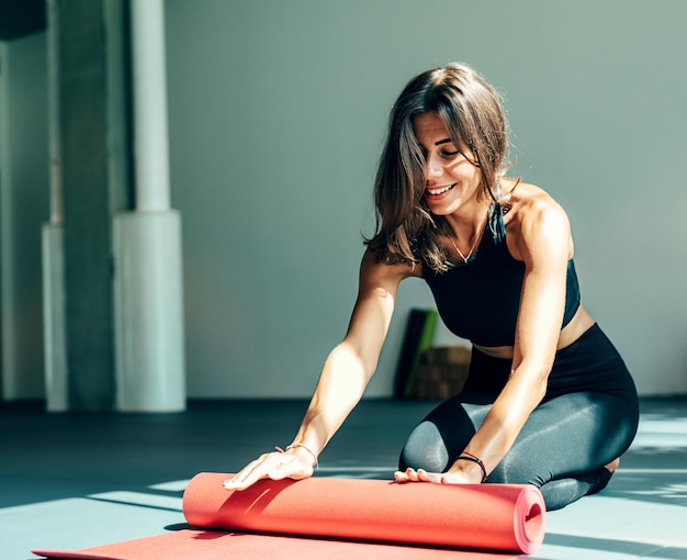 Foto grátis mulher sorridente em roupas esportivas rolando tapete no estúdio de ioga