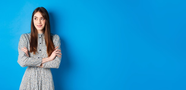 Foto grátis mulher sorridente em vestido romântico cruzando os braços no peito e olhando para a esquerda no logotipo em pé contra azul b