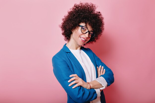 Mulher sorridente engraçada com cachos usando óculos e poses de jaqueta azul rosa