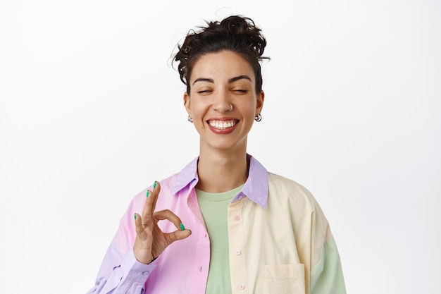 Foto grátis mulher sorridente feliz fecha os olhos, mostrando sinal de ok, gesto zero, sem problemas, elogia uma coisa boa incrível, de pé com uma roupa elegante contra um fundo branco.