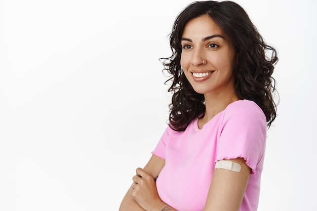 Mulher sorridente feliz foi vacinada contra SARS COV covid-19, desviando o olhar com os braços cruzados, gesso médico no ombro onde a vacina foi feita, fundo branco