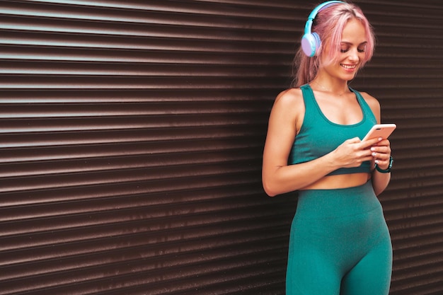 Mulher sorridente fitness em roupas esportivas verdes com cabelo rosa Jovem modelo bonito com corpo perfeitoFêmea na rua perto da parede da persiana Ouvir música em fones de ouvido sem fioUsando aplicativos