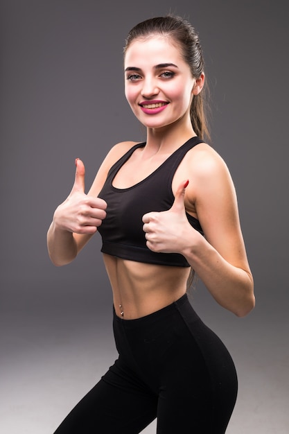 Mulher sorridente fitness mostrando o polegar para cima o sinal sobre parede cinza