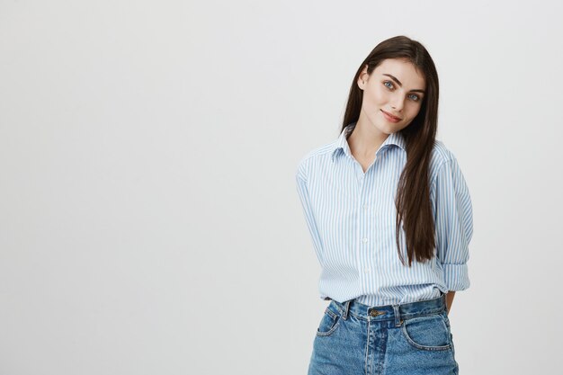 Mulher sorridente fofa de camiseta e jeans de mãos dadas atrás das costas e cabeça inclinada