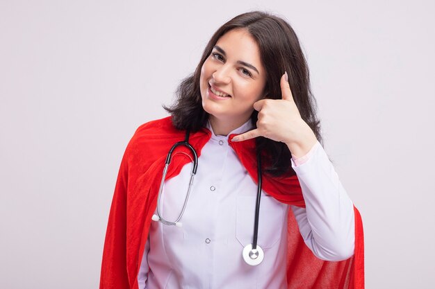 Mulher sorridente jovem super-herói vestindo uniforme de médico e estetoscópio olhando para frente fazendo um gesto de chamada isolado na parede