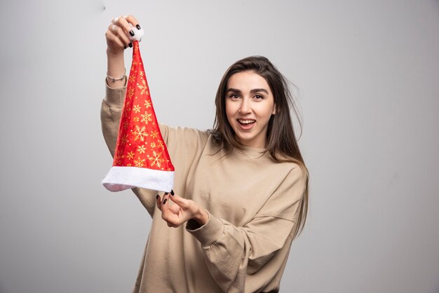 Mulher sorridente mostrando o chapéu de Papai Noel em um fundo cinza.