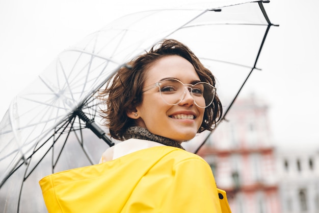 Mulher sorridente na capa de chuva amarela e óculos, tendo prazer em caminhar pela cidade sob o grande guarda-chuva transparente durante o dia chuvoso frio