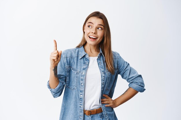 Mulher sorridente pensativa com cabelo loiro, levantando o dedo e apontando para cima, tendo um bom argumento ou ideia, mostrando um anúncio, em pé no branco