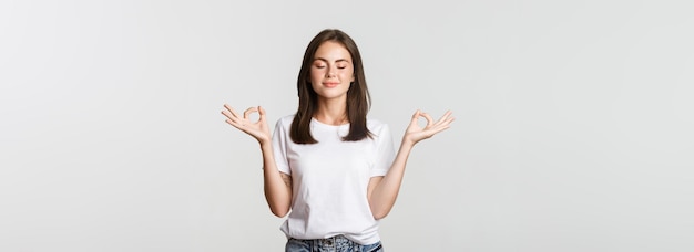 Mulher sorridente relaxada fecha os olhos e medita fazendo yoga em pé fundo branco