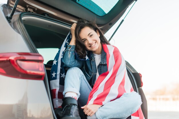 Mulher sorridente segurando bandeira grande EUA na mala do carro