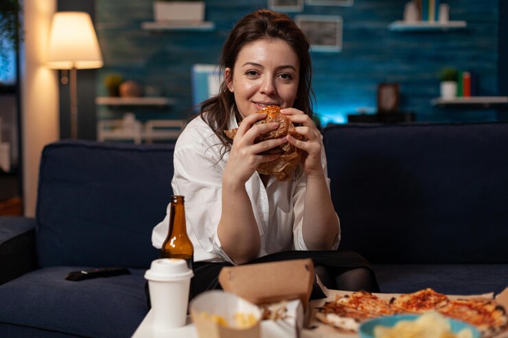 Foto grátis mulher sorridente segurando hambúrguer saboroso nas mãos, olhando para a comédia de comédia de televisão em pé no sofá em frente à mesa com o menu de fast food para viagem. pessoa feliz assistindo programa de tv com jantar para viagem.