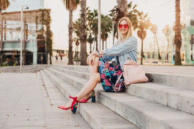 mulher sorridente sentada na escada em uma rua da cidade com saia estampada elegante e jaqueta jeans grande demais com mochila de couro e óculos de sol rosa