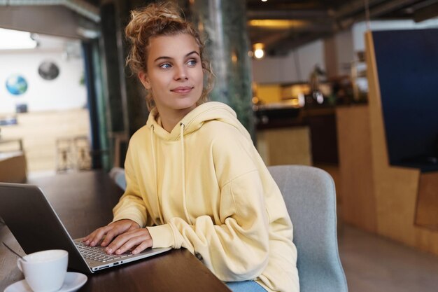 Mulher sorridente sentada no café com laptop olhando para fora da janela satisfeita Freelancer feminino criativo obtendo inspiração trabalhando ao ar livre na cafeteria Menina estudando on-line no café com laptop