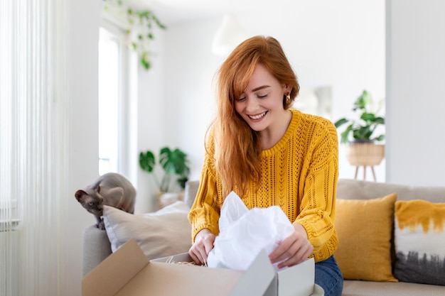 Mulher sorridente sentada no sofá em casa pacote de correio aberto compras on-line comprando mercadorias na internet cliente jovem feliz desembalar pacote de envio postal satisfeito com pedido ou entrega