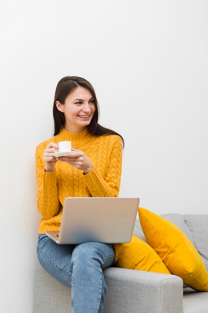 Mulher sorridente, sentado no sofá ao lado do laptop e segurando a xícara de café