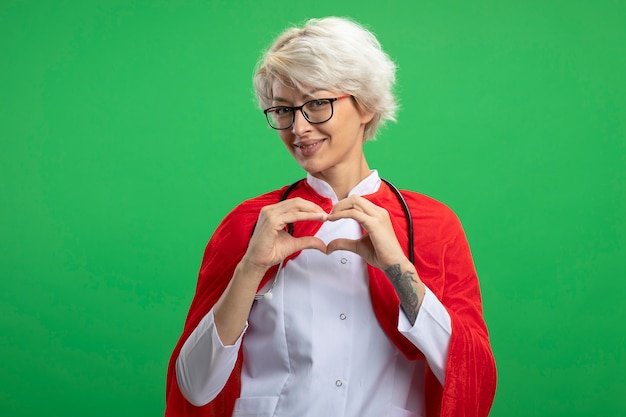 Mulher sorridente super-heroína eslava em uniforme de médico com capa vermelha e estetoscópio em óculos ópticos gestos coração isolado na parede verde com espaço de cópia