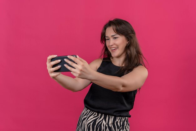 Mulher sorridente usando camiseta preta tira uma selfie na parede rosa