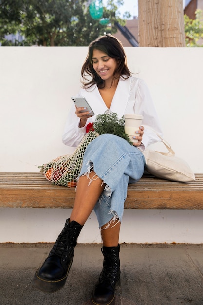 Mulher sorridente usando seu smartphone ao ar livre enquanto toma um café