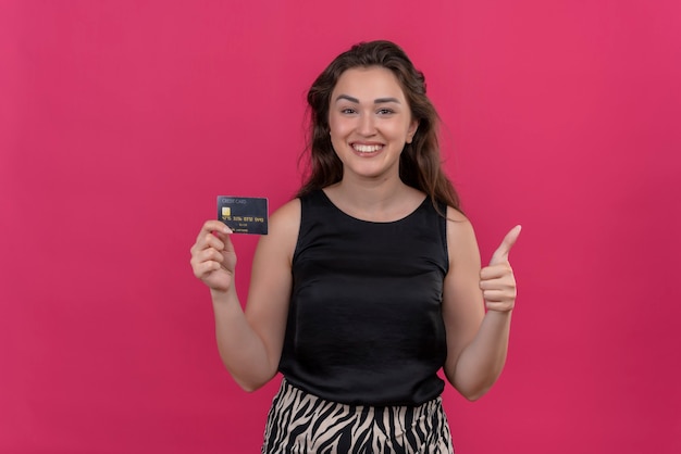 Mulher sorridente, vestindo camiseta preta, segurando um cartão do banco e apontando para ela na parede rosa