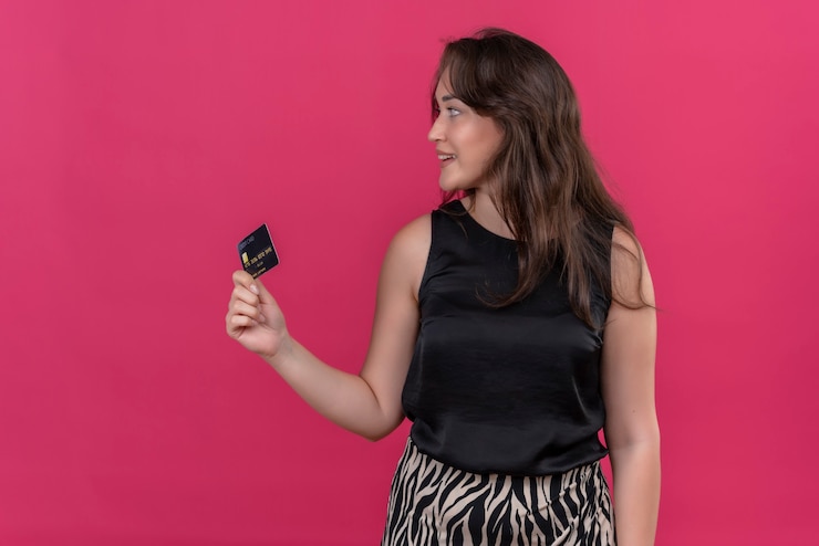 Foto grátis mulher sorridente, vestindo camiseta preta, segurando um cartão do banco e olhando para o lado na parede rosa