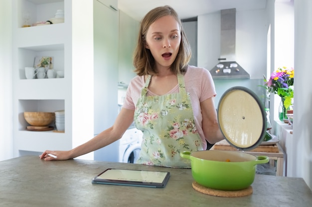 Mulher surpreendida com a boca aberta, olhando para uma panela na cozinha dela, usando o tablet no balcão. vista frontal. cozinhar em casa conceito