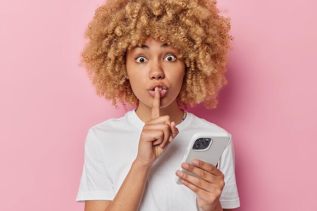 Mulher surpreendida com cabelo espesso encaracolado mostra gesto de silêncio mantém o dedo indicador sobre os lábios usa celular usa camiseta branca casual isolada sobre fundo rosa Não conte a ninguém sobre isso