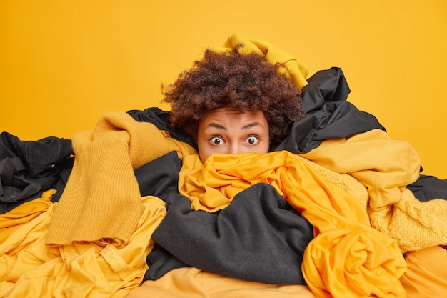 Foto grátis mulher surpreendida com cabelos cacheados olha chocada enterrada em uma grande pilha de roupas amarelas e pretas faz limpeza de guarda-roupa coleta roupas para lavar