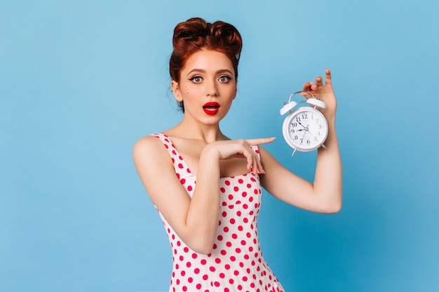 Mulher surpreendida com maquiagem brilhante, mostrando o tempo. Foto de estúdio de garota Pin-up chocada segurando o relógio no espaço azul.