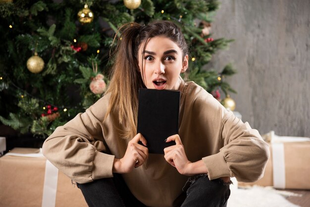 Mulher surpreendida sentada com um caderno preto perto da árvore de Natal.