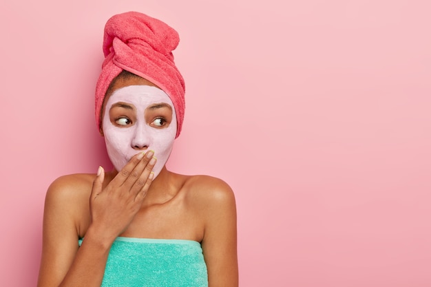 Mulher surpresa cobre a boca, aplica máscara nutritiva para remover células mortas, usa toalha enrolada na cabeça, fica de pé contra a parede rosa