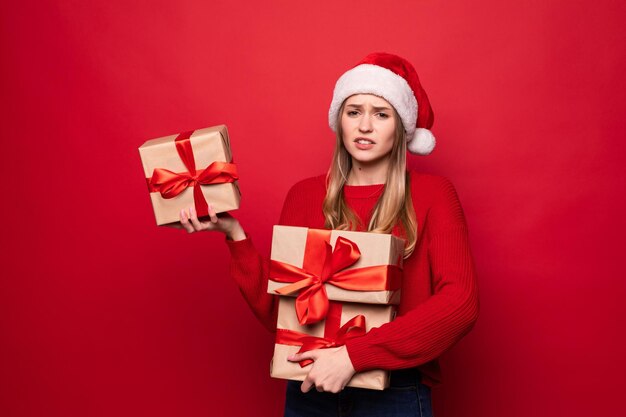 Mulher surpresa e animada com roupa vermelha de Papai Noel segurando uma pilha de presentes isolados na parede vermelha