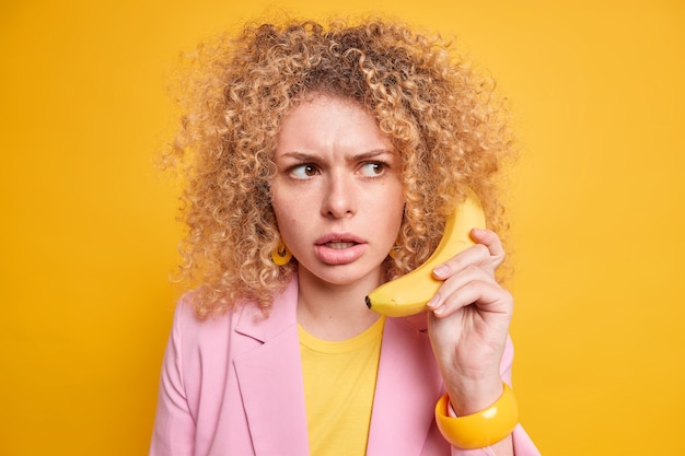 Mulher tem expressão estrita zangada desempenha o papel de chefe mantém banana perto da orelha finge estar atendendo a uma ligação telefônica vestida com uma jaqueta estilosa