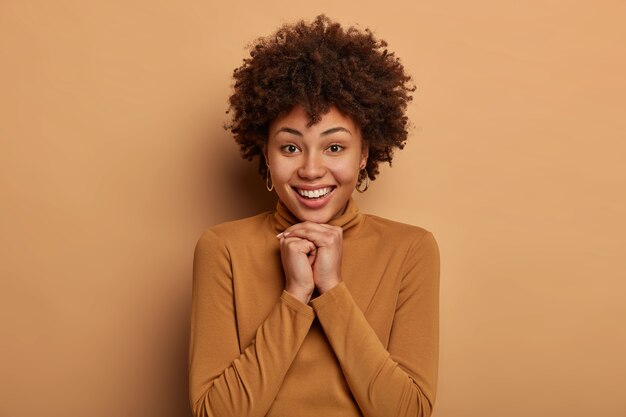 Mulher tenra e feminina com penteado encaracolado, olha sincero para a câmera, mantém as mãos juntas sob o queixo e usa uma blusa de gola alta marrom