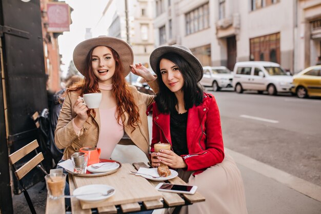 Mulher tímida de saia bege posando com prazer em um café ao ar livre durante o almoço com um amigo