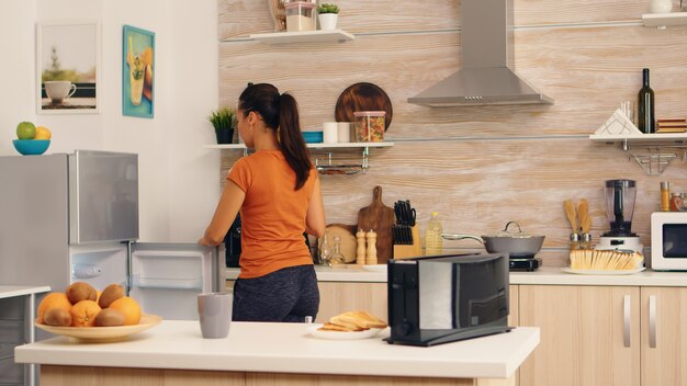 Mulher tirando ovos da geladeira pela manhã no café da manhã. Dona de casa pegando ovos saudáveis e outros ingredientes da geladeira em sua cozinha.