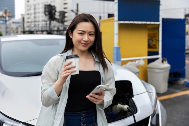 Mulher tomando café enquanto seu carro elétrico está carregando e usando smartphone