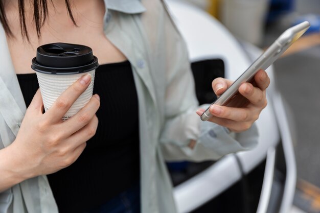 Mulher tomando café enquanto seu carro elétrico está carregando e usando smartphone