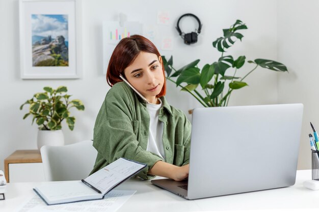 Mulher trabalhando remotamente em casa com um laptop. jovem empresária falando ao telefone