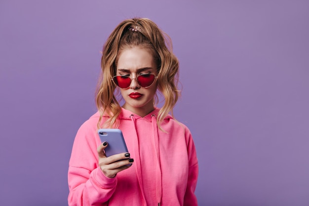 Foto grátis mulher triste com capuz e óculos de sol vermelhos lê mensagem em seu telefone retrato de mulher de mau humor segurando celular em fundo roxo isolado