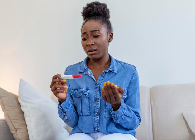 Mulher triste solteira reclamando segurando um teste de gravidez sentada em um sofá na sala de estar em casa Garota negra deprimida segurando teste de gravidez negativo