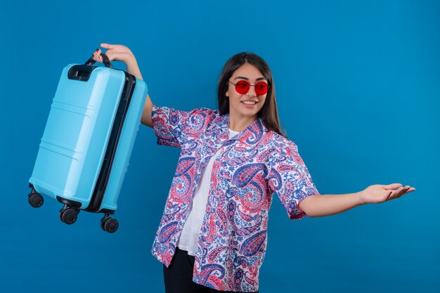 mulher turista usando óculos escuros vermelhos segurando uma mala de viagem sorrindo amigável em pé com os braços abertos em um gesto de boas-vindas no azul isolado