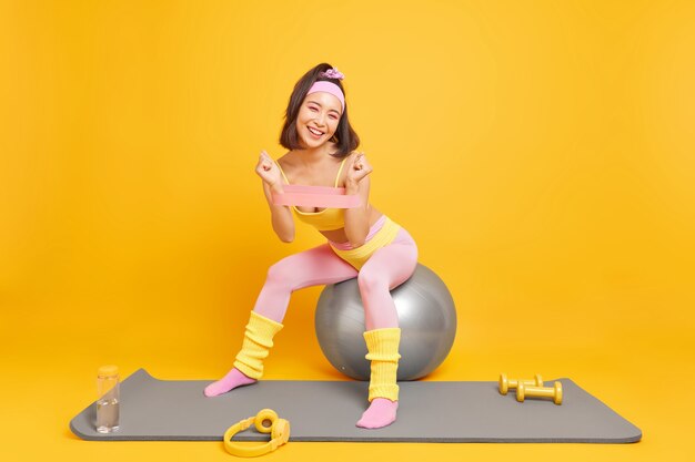 Mulher usa acessórios esportivos treina os músculos dos braços com banda de resistência senta na bola de fitness vestida com roupas esportivas