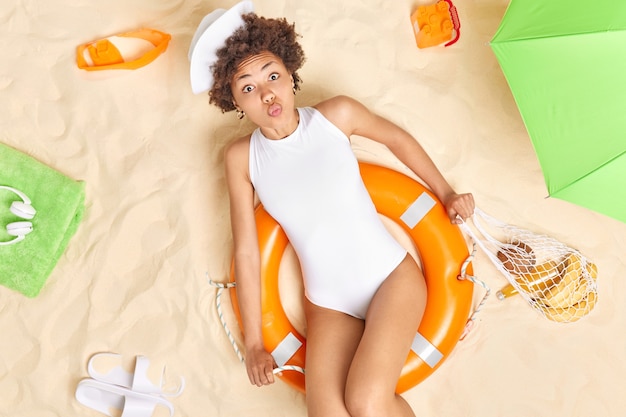 Mulher usa chapéu de sol branco e biquíni sentada em bóia salva-vidas inflada mantém os lábios arredondados quer beijar alguém gosta de viajar poses de férias na praia segura bolsa de rede com frutas