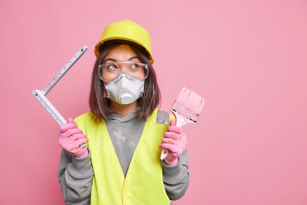 Mulher usa óculos de segurança, respirador e capacete segura escova de pintura fita métrica conserta suportes de apartamentos