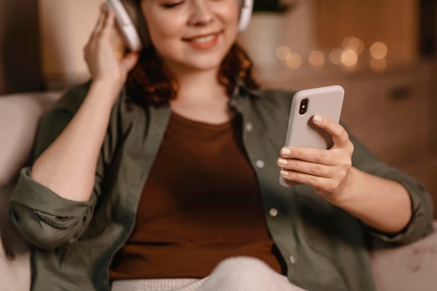 Mulher usando fones de ouvido modernos e smartphone em casa no sofá