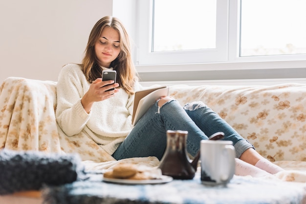 Mulher, usando, smartphone, ligado, sofá, perto, tabela, com, bebida, e, biscoitos